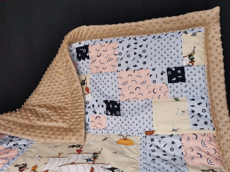 Peut inclure: Une couverture b&eacute;b&eacute; beige avec un motif patchwork sur le th&egrave;me nautique. La couverture est faite d'un tissu doux et pelucheux et a une texture matelass&eacute;e. Le motif patchwork comprend divers motifs, tels que des ancres, des bateaux et des &eacute;toiles. La couverture est parfaite pour c&acirc;liner et garder les b&eacute;b&eacute;s au chaud.