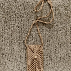 May include: A beige crochet phone bag with a long strap. The bag features a chevron pattern and a button closure. The strap is also crocheted. The bag is made of a soft, textured yarn.