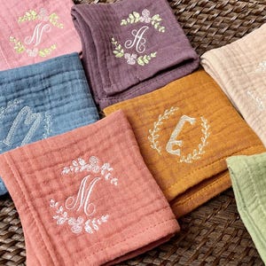 May include: Seven embroidered cotton muslin squares in various colors, each with a different letter monogram in a floral wreath. The colors include light blue, pink, purple, orange, beige, green, and red.