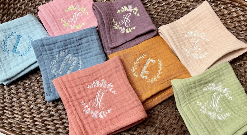 May include: Seven embroidered cotton muslin squares in various colors, each with a different letter monogram in a floral wreath. The colors include light blue, pink, purple, orange, beige, green, and red.