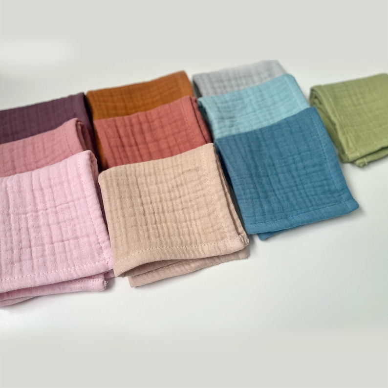 May include: A set of ten square muslin cloths in various colors, including pink, beige, brown, red, blue, green, and gray. The cloths are folded and arranged in a row on a white surface.