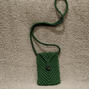 May include: A handmade, dark green crochet phone bag with a long strap. The bag features a chevron pattern and a button closure. The strap is also crocheted. The bag is shown on a textured, light gray surface.
