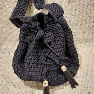 May include: A navy blue crochet drawstring bag with a long strap. The bag features a rounded shape and is adorned with two tassels with gold-colored caps. The bag is made of a thick, textured yarn.