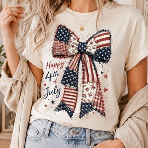 May include: A cream-coloured t-shirt featuring a large, frayed-edge bow graphic with a patchwork design of American flag patterns, stars, and stripes. The text "Happy 4th of July" is printed in red and blue script next to the bow graphic.