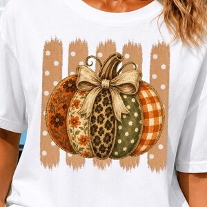 May include: A white short-sleeve t-shirt featuring a central graphic of a pumpkin with a tan bow. The pumpkin is divided into segments with floral, leopard, polka dot, and plaid patterns, set against a background of vertical tan brushstrokes.