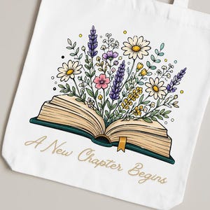 May include: A white canvas tote bag featuring a colorful illustration of an open book with a bouquet of daisies, lavender, and yellow wildflowers emerging from the pages. The text A New Chapter Begins is printed in gold cursive below the book.