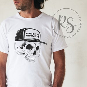 May include: White t-shirt featuring a black and white skull graphic wearing a baseball cap. The cap has the text "SON OF A SINNER" on the front. The design is a detailed illustration, suitable for those interested in gothic or edgy apparel.