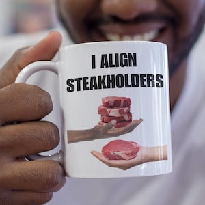 Peut inclure: Une tasse à café blanche avec un graphique noir et blanc d'une main tenant une pile de steaks. Le texte sur la tasse est "I align steakholders".