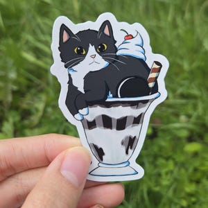 Puede incluir: Una pegatina con un gato blanco y negro descansando en un vaso de helado. El helado tiene salsa de chocolate, crema batida, una cereza y una barquilla. El gato tiene ojos amarillos y una mancha blanca en el pecho.