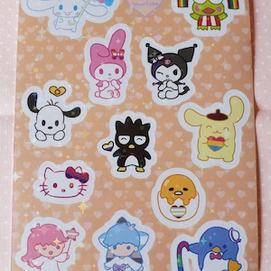 Puede incluir: Una hoja de pegatinas Sanrio Pride con personajes como Hello Kitty, My Melody, Keroppi y Badtz-Maru. Cada personaje sostiene un corazón con la bandera del orgullo.
