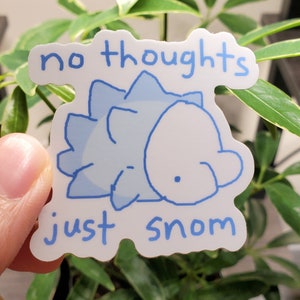 May include: A white sticker featuring a light blue cartoon Snom character and the text "no thoughts just snom" in light blue. The sticker is held in front of a green plant.
