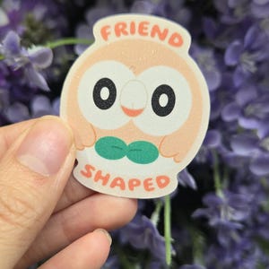 May include: A sticker with a cartoon owl design. The owl is peach-colored with large black eyes, a green bow tie, and the words "FRIEND SHAPED" in orange. The sticker is held against a background of purple flowers.
