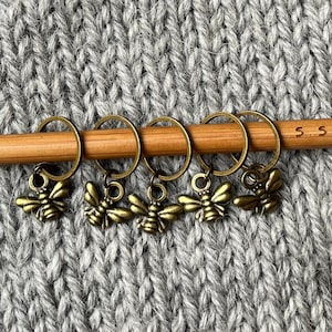 Könnte beinhalten: Fünf goldene Bienen-Maschenmarkierer auf einer Holzstricknadel mit der Gravur "5,5 mm" auf der Nadel. Die Maschenmarkierer befinden sich auf einem grauen Strickstoff.