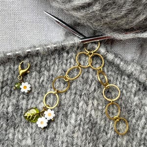 Könnte beinhalten: Nahaufnahme eines grauen Strickstoffes mit zwei stichförmigen Stichmarkierungen und einer goldenen Ketten-Stichmarkierung. Die Stichmarkierungen sind mit einer kleinen Schlaufe am Stoff befestigt.