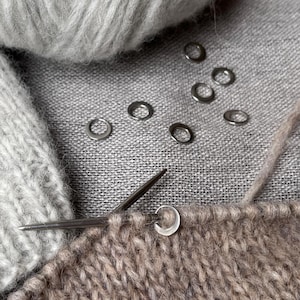 May include: A light grey ball of yarn sits next to a partially knitted piece of light brown fabric on a needle. Several small, circular silver stitch markers are scattered across the textured grey fabric surface near the knitting project.