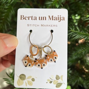 Könnte beinhalten: Drei fuchsförmige Maschenmarkierer mit orangefarbenen, weißen und schwarzen Perlendetails. Die Markierer sind an goldenen Ringen befestigt und werden auf einer weißen Karte mit dem Text "Berta un Maija STITCH MARKERS" präsentiert.
