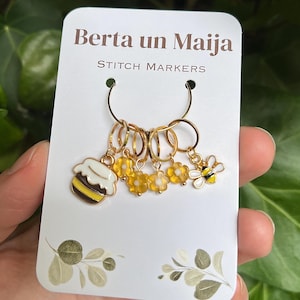 Könnte beinhalten: Goldene Maschenmarkierer mit Bienen- und Blumenanhängern. Die Anhänger umfassen einen Cupcake, gelbe Blumen und eine Biene. Auf der Karte steht "Berta un Maija STITCH MARKERS".