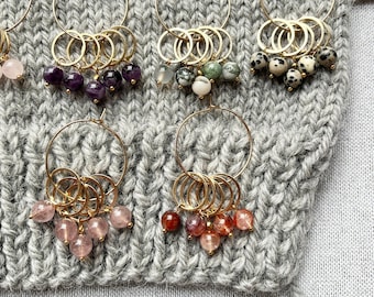 Puede incluir: Una colección de pendientes de aro dorados, cada uno adornado con un grupo de cuentas de piedras preciosas de colores. Las cuentas varían en color, incluyendo rosa, verde, negro y patrones moteados. Los pendientes se exhiben sobre una tela de punto gris.