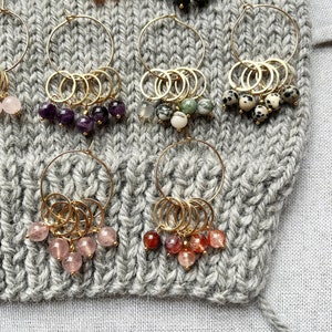 Könnte beinhalten: Eine Sammlung goldfarbener Creolen, jede mit einem Cluster aus farbigen Edelsteinperlen verziert. Die Perlen variieren in der Farbe, darunter rosa, grün, schwarz und gesprenkelte Muster. Die Ohrringe werden auf einem grauen Strickstoff präsentiert.