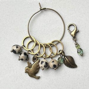 Könnte beinhalten: Ein goldfarbener Metallring mit einer Reihe kleinerer Ringe, die daran befestigt sind, und einem Anhänger mit einem Vogel- und Blattmotiv. Der Ring ist mit schwarz-weißen, gefleckten Perlen verziert.
