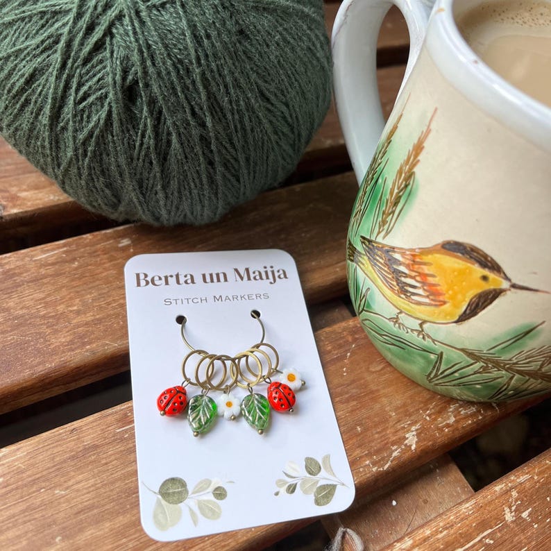 K&ouml;nnte beinhalten: Ein Satz goldener Maschenmarkierer mit roten Marienk&auml;fer- und wei&szlig;en Blumenanh&auml;ngern. Die Markierer befinden sich auf einer wei&szlig;en Karte mit dem Text "Berta un Maija Stitch Markers".
