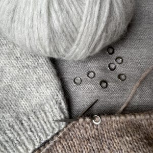 May include: A light grey ball of yarn, a knitted light grey fabric piece, a brown knitted fabric piece on a metal knitting needle, and six small silver-coloured metal stitch markers arranged in a cluster on a grey textile surface.