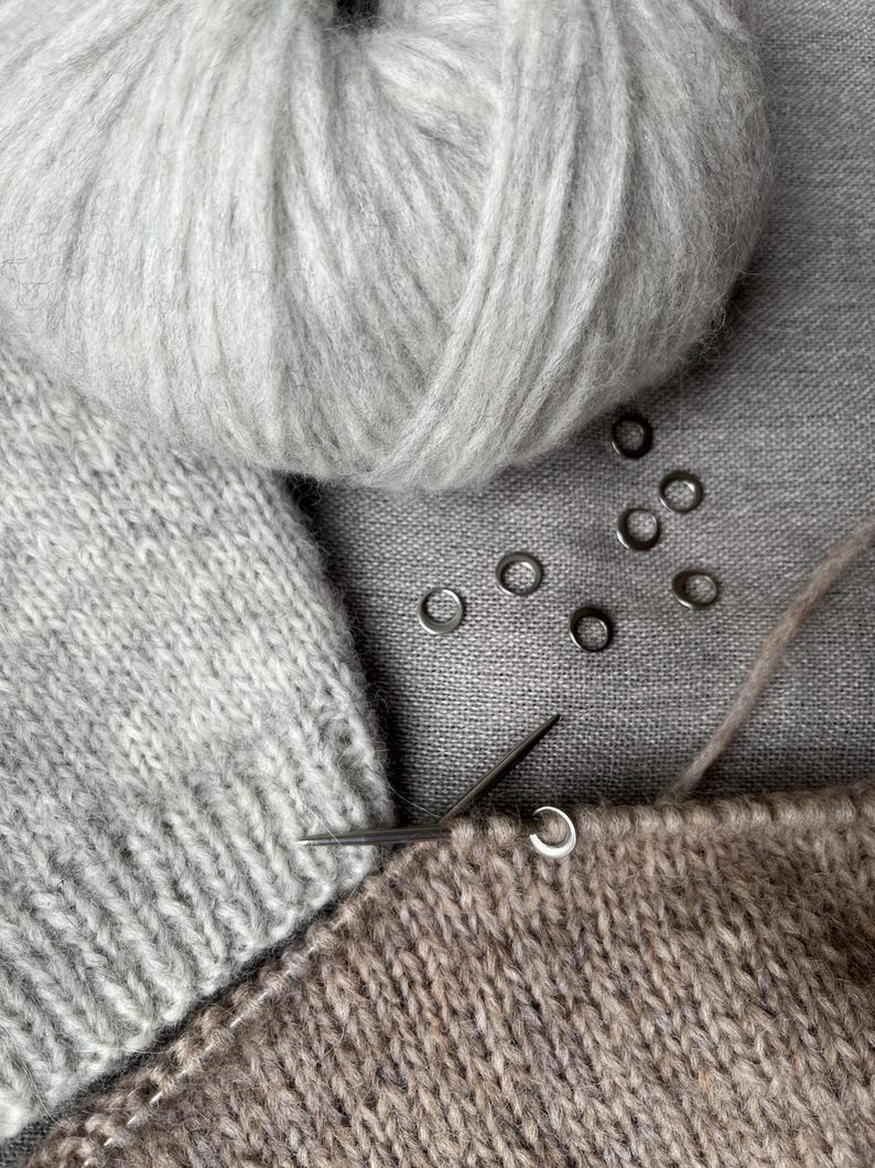 May include: A light grey ball of yarn, a knitted light grey fabric piece, a brown knitted fabric piece on a metal knitting needle, and six small silver-coloured metal stitch markers arranged in a cluster on a grey textile surface.