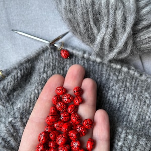 K&ouml;nnte beinhalten: Eine Hand h&auml;lt einen Haufen roter Marienk&auml;fer-Perlen. Die Perlen sind auf der Handfl&auml;che verteilt und in einem zuf&auml;lligen Muster angeordnet. Die Perlen sind aus einem glatten, gl&auml;nzenden Material gefertigt und haben schwarze Punkte. Die Perlen sind von einem grauen Strickstoff umgeben.
