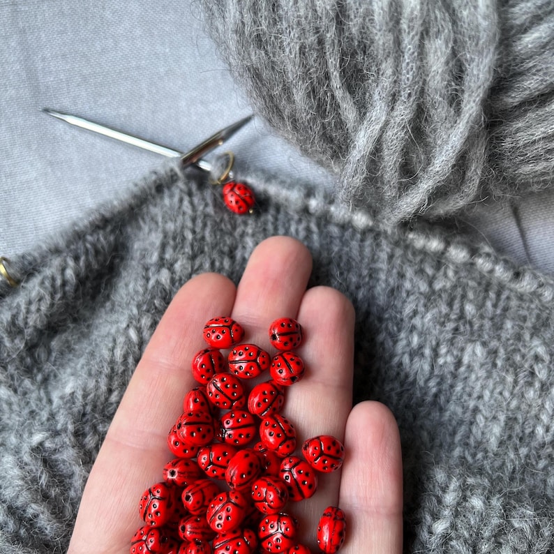 K&ouml;nnte beinhalten: Eine Hand h&auml;lt einen Haufen roter Marienk&auml;fer-Perlen. Die Perlen sind auf der Handfl&auml;che verteilt und in einem zuf&auml;lligen Muster angeordnet. Die Perlen sind aus einem glatten, gl&auml;nzenden Material gefertigt und haben schwarze Punkte. Die Perlen sind von einem grauen Strickstoff umgeben.