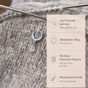 May include: A silver-toned stainless steel stitch marker ring featuring a clear faceted glass bead charm. The text on the right side lists features including Aus Edelstahl gefertigt, Abnehmbarer Ring, Mit klarer b&ouml;hmischer Glasperle, and Minimalistisch & leicht.