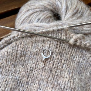 May include: A silver-toned metal stitch marker with a small, clear, faceted bead charm attached to a circular clasp. The marker is hooked into a piece of light brown, fuzzy knitted fabric, which is resting behind a long, thin, metallic knitting needle.