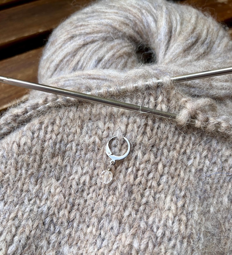 May include: A silver-toned metal stitch marker with a small, clear, faceted bead charm attached to a circular clasp. The marker is hooked into a piece of light brown, fuzzy knitted fabric, which is resting behind a long, thin, metallic knitting needle.