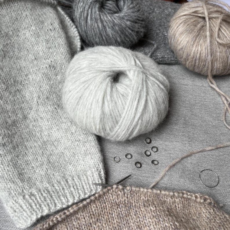 May include: Three balls of yarn in shades of light grey, dark grey, and beige are arranged with a partially knitted light grey garment and a partially knitted beige garment. Several small metal stitch markers are scattered on the fabric.