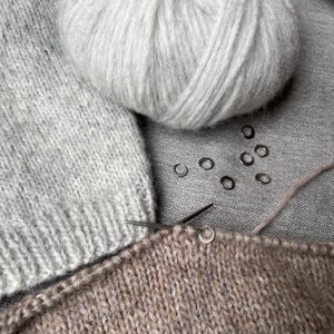 May include: A light grey ball of yarn sits next to a piece of light grey knitted fabric and a piece of tan knitted fabric. A thin metal knitting needle is inserted into the tan fabric, which features several small silver stitch markers.