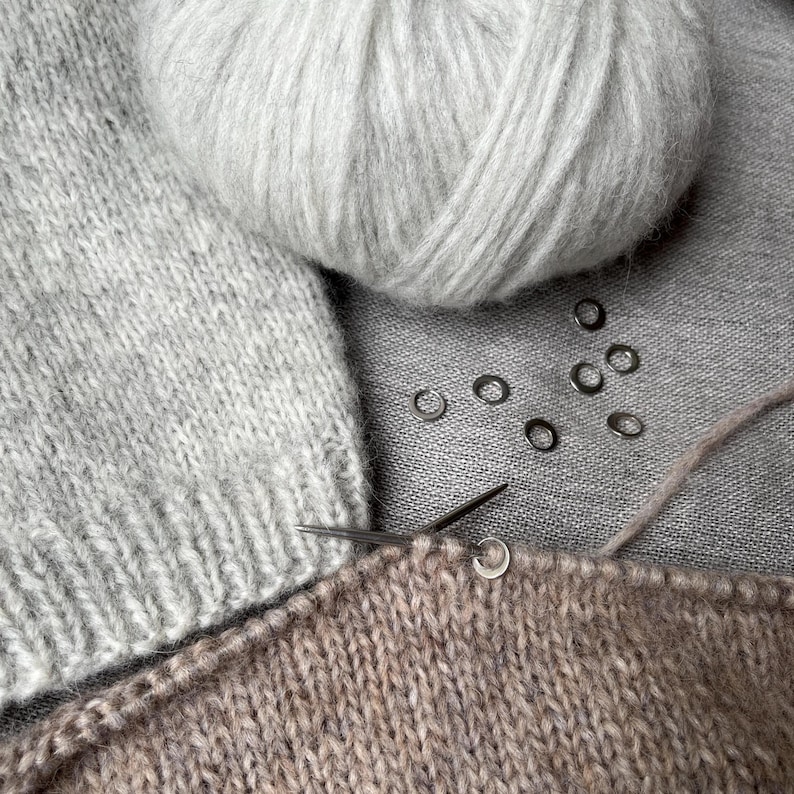 May include: A light grey ball of yarn sits next to a piece of light grey knitted fabric and a piece of tan knitted fabric. A thin metal knitting needle is inserted into the tan fabric, which features several small silver stitch markers.