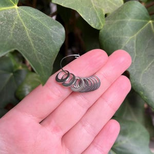 May include: A set of small, circular, silver-toned metal stitch markers arranged on a thin, flexible wire hoop. The markers are smooth, uniform in size, and feature a simple, open ring design, providing a functional tool for knitting or crochet projects.