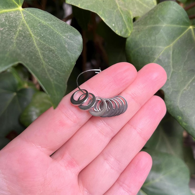 May include: A set of small, circular, silver-toned metal stitch markers arranged on a thin, flexible wire hoop. The markers are smooth, uniform in size, and feature a simple, open ring design, providing a functional tool for knitting or crochet projects.