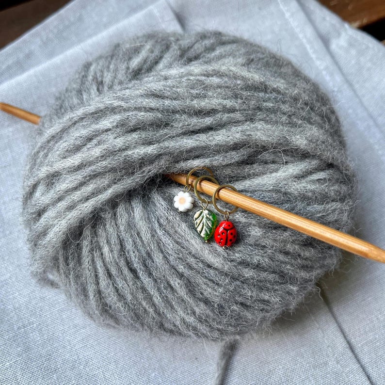 K&ouml;nnte beinhalten: Eine Kugel graues Garn mit einer Stricknadel aus Holz und drei Maschenmarkierern. Die Maschenmarker sind eine wei&szlig;e Blume, ein gr&uuml;nes Blatt und ein roter Marienk&auml;fer.