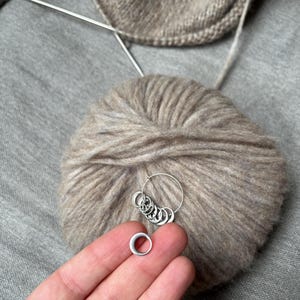 May include: A set of silver-toned metal stitch markers for knitting, featuring a circular ring design. One marker is held separately, while the remaining markers are organised on a larger silver-toned metal ring, resting against a ball of beige yarn.