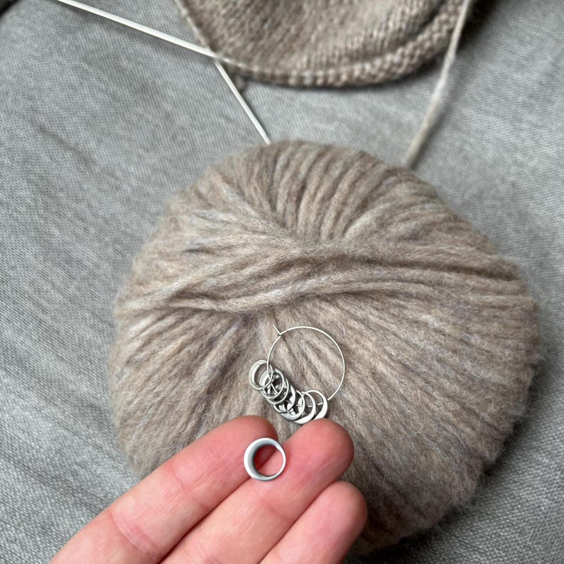 May include: A set of silver-toned metal stitch markers for knitting, featuring a circular ring design. One marker is held separately, while the remaining markers are organised on a larger silver-toned metal ring, resting against a ball of beige yarn.