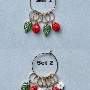K&ouml;nnte beinhalten: Zwei S&auml;tze Stichmarker mit einem Reifen, Ringen und Charms. Set 1 hat gr&uuml;ne Bl&auml;tter und rote Marienk&auml;fer. Set 2 hat gr&uuml;ne Bl&auml;tter, rote Marienk&auml;fer und wei&szlig;e Blumen. Set 1 und Set 2 sind beschriftet.