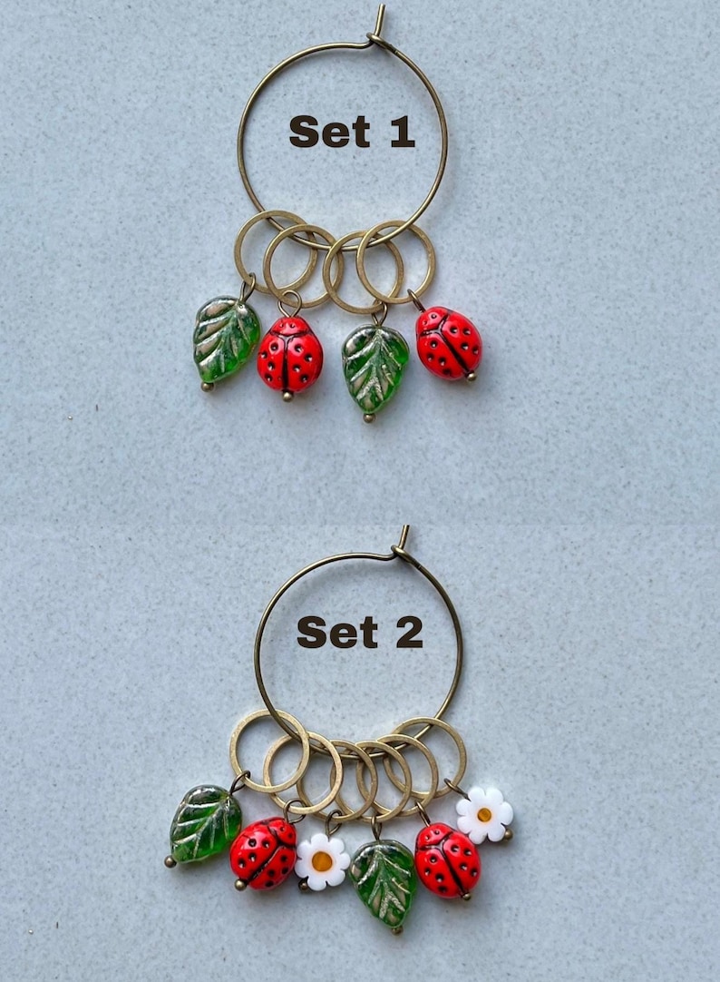 K&ouml;nnte beinhalten: Zwei S&auml;tze Stichmarker mit einem Reifen, Ringen und Charms. Set 1 hat gr&uuml;ne Bl&auml;tter und rote Marienk&auml;fer. Set 2 hat gr&uuml;ne Bl&auml;tter, rote Marienk&auml;fer und wei&szlig;e Blumen. Set 1 und Set 2 sind beschriftet.
