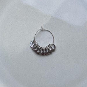 May include: A silver-toned metal stitch marker ring featuring a thin circular wire hoop adorned with a series of nine smaller, crescent-shaped metal charms that are attached to the main hoop and hang in a curved arrangement along the bottom.