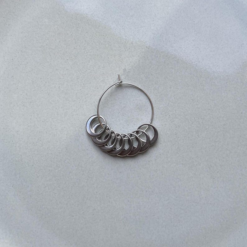 May include: A silver-toned metal stitch marker ring featuring a thin circular wire hoop adorned with a series of nine smaller, crescent-shaped metal charms that are attached to the main hoop and hang in a curved arrangement along the bottom.