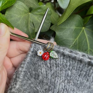 K&ouml;nnte beinhalten: Nahaufnahme eines grauen Strickstoffes mit einem Metall-Maschenmarkierer mit einem Marienk&auml;fer-, Blatt- und Blumenanh&auml;nger. Der Maschenmarkierer befindet sich auf einer Stricknadel.