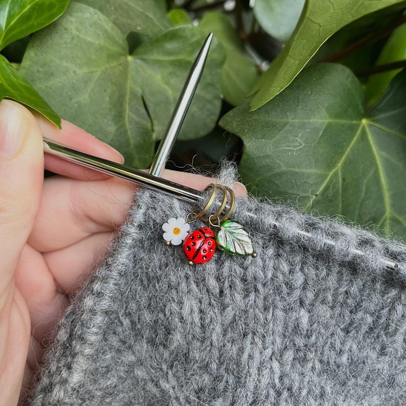 K&ouml;nnte beinhalten: Nahaufnahme eines grauen Strickstoffes mit einem Metall-Maschenmarkierer mit einem Marienk&auml;fer-, Blatt- und Blumenanh&auml;nger. Der Maschenmarkierer befindet sich auf einer Stricknadel.