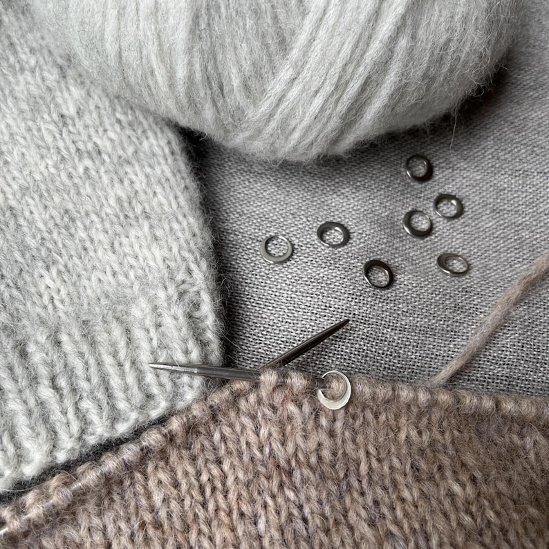 May include: A light grey ball of yarn sits above a piece of light brown knitted fabric on a knitting needle. Several small, circular silver stitch markers are scattered across the fabric, with one marker attached to the needle.