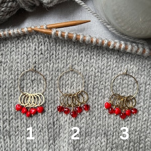 Op de afbeelding: Drie gouden steekmarkeerders met rode hartcharms. De steekmarkeerders zijn genummerd 1, 2 en 3.