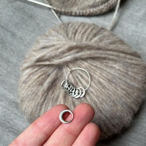 May include: A set of silver-coloured metal stitch markers consisting of a larger circular ring holding several smaller, thinner circular rings. The markers are positioned against a ball of light brown, fuzzy yarn with knitting needles visible.