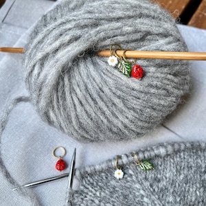K&ouml;nnte beinhalten: Graue Garnkugel mit einer Stricknadel aus Holz und drei Maschenmarkern. Die Maschenmarker sind ein Marienk&auml;fer, eine Blume und ein Blatt. Das Garn liegt auf einer grauen Oberfl&auml;che.
